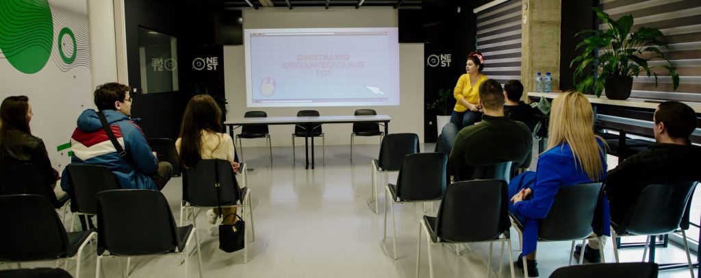 Mala grupa ljudi sjedi u modernoj učionici okrenuta ka platnu na kojem se prikazuje prezentacija pod nazivom „Digitalno organizovanje 101“. Žena u žutom džemperu Vesna Jusup stoji i obraća se publici koja sjedi na crnim stolicama.
